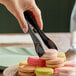 A pair of black disposable plastic tongs being used to pick up a macaron from a stack of assorted macarons.