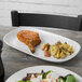 A piece of meat on a Tuxton rectangular white china platter on a table with a salad and a drink.