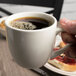 A hand holding a Tuxton white china coffee mug filled with coffee.