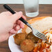 A hand holding a WNA Comet Reflections Duet stainless steel look plastic fork with a black handle over a plate of food.