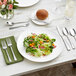 An Acopa stainless steel butter knife on a green napkin next to a white plate with salad