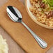 An Acopa Edgeworth stainless steel serving spoon next to a bowl of rice with mushrooms and green sprouts on a table.