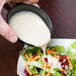 A person pouring a black Solo sauce cup of white sauce onto a salad.