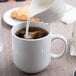 A white pitcher pouring milk into a white Libbey stacking mug with coffee.