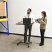 A man standing at a Victor High Rise adjustable height desk holding a laptop and a woman reading.