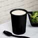 A black Tablecraft salad dressing crock filled with white salad dressing on a table with salad and a black spoon.