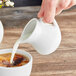 A hand pouring milk from an Acopa white creamer into a cup of coffee.
