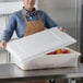 A woman wearing a brown apron holding a white Choice food storage container filled with apples.