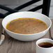 A Schonwald white porcelain bowl filled with soup and a spoon on a white plate with a cup of coffee.