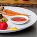 A white plate of food with a sauce and vegetables with a white round sauce cup on it.