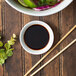 A table with a bowl of food and a white porcelain round sauce cup filled with soy sauce next to chopsticks.