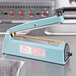 An Omcan manual impulse bag sealer on a counter with a blue handle.