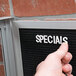 A person pointing at a black Aarco enclosed message board with a white felt sign inside.