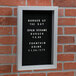 An Aarco black felt message board with white text that says "Burger of the Day" on a brick wall.