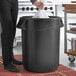 A man in a professional kitchen putting a plastic bag into a black Rubbermaid BRUTE trash can.