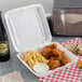 A Dart white foam take out container with chicken and fries next to a bottle of beer.