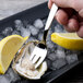 A hand holding a Chef & Sommelier stainless steel cocktail fork over an oyster on ice with a lemon wedge.