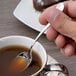 A person holding a Chef & Sommelier stainless steel demitasse spoon over a cup of liquid.