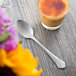 A Libbey stainless steel teaspoon next to a glass of liquid on a wood table.