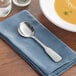 An Acopa stainless steel bouillon spoon on a blue napkin next to a bowl of soup.