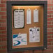 An Aarco bronze indoor lighted bulletin board cabinet with cork papers attached to it.