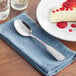 An Acopa stainless steel spoon on a blue napkin next to a plate of cheesecake.