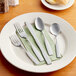 A Choice Delmont stainless steel iced tea spoon on a table with other silverware.