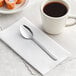 A Choice Windsor stainless steel teaspoon on a napkin next to a cup of coffee.