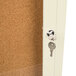 An ivory indoor bulletin board cabinet with a cork board inside and a key in the door lock.