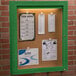 An Aarco green indoor lighted bulletin board cabinet with cork board inside and a green frame.