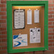 An Aarco green indoor lighted bulletin board cabinet with cork board inside and papers attached to it.