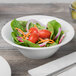 A white Schonwald porcelain bowl filled with a salad of tomatoes, spinach, and onions on a wooden table.