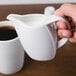 A person holding a Schonwald white porcelain creamer pouring milk into a cup of coffee.