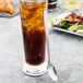 A glass of brown liquid with ice and a Libbey stainless steel iced tea spoon on a table.