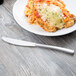 A Libbey stainless steel dinner knife with a serrated blade on a wood surface next to a plate of food.