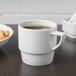 A white Schonwald stackable mug filled with brown liquid on a table with a bowl of food.