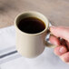 A person's hand holding a Thunder Group Nustone Sand bulbous mug filled with coffee.