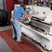 A man in a blue shirt and jeans stands on a red Cactus Mat VIP Prima floor mat while working on a machine.