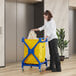 A woman pushing a blue and yellow Lavex Commercial laundry cart with a white bag.