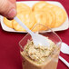 A hand holding a Fineline white plastic tasting spoon over a container of food.