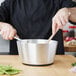 A chef using a Vollrath Wear-Ever aluminum sauce pan with a wooden stick to stir vegetables on a stove.