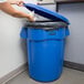 A woman putting a plastic bag into a blue Rubbermaid BRUTE trash can.