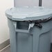 A Rubbermaid BRUTE grey trash can with a black lid in a corporate office cafeteria.