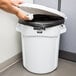 A hand putting a plastic bag into a white Rubbermaid BRUTE trash can.