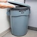 A woman holding a garbage bag and putting it in a Rubbermaid BRUTE grey trash can in a corporate office cafeteria.