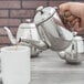A hand pouring coffee into a Vollrath stainless steel teapot.