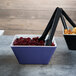 A blue and white square melamine bowl filled with cranberries on a table with a bowl of food and tongs.