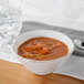A white Urban Mill melamine bowl filled with soup and vegetables next to a glass of water.