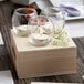 A white Leola tea light candle in a glass on a wooden table with two other candles in glasses.