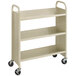 A beige metal three-shelf book cart with black wheels.